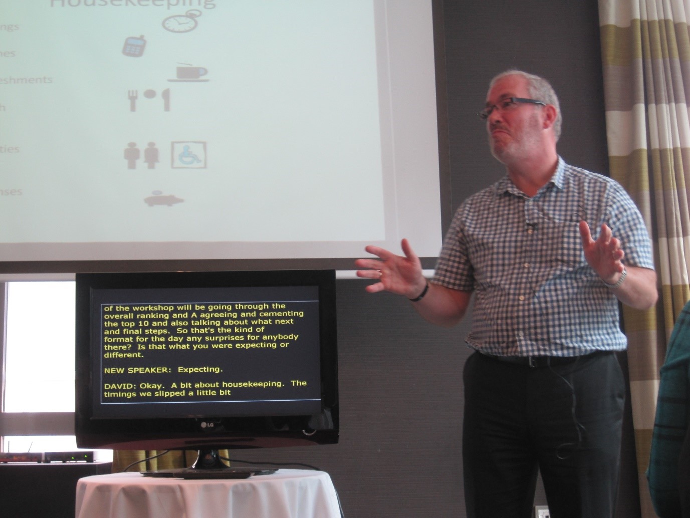 A man speaking at a conference with real-time speech-to-text reporting on a screen beside him
